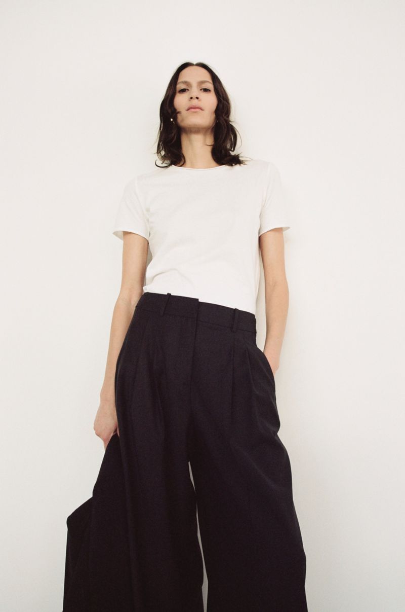Brunette woman wearing a white t-shirt and black wide-leg trousers poses against a plain white wall in a fashion shot.