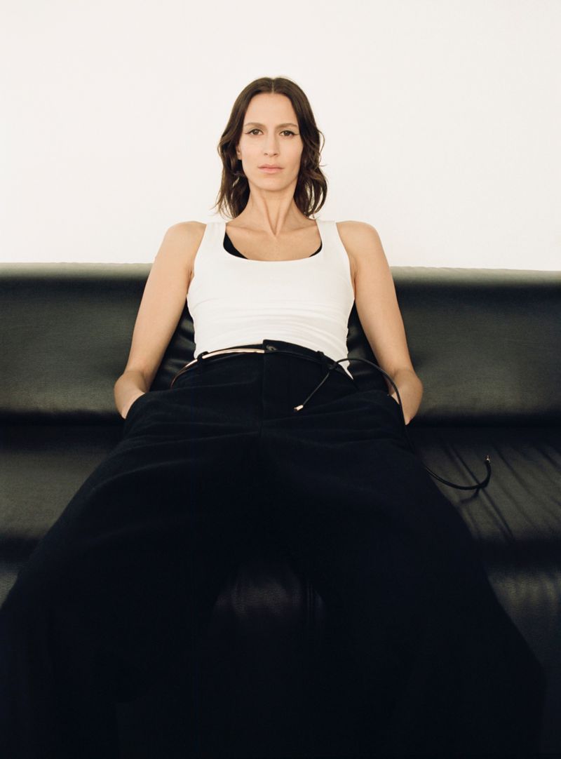 A woman in a white tank top and black pants is sitting on a black couch in front of a white wall looking directly at the camera.