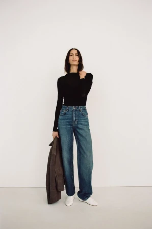 Model Jeanne Cadieu poses against a white studio backdrop wearing a black long-sleeve top, wide-leg blue jeans, and white shoes.