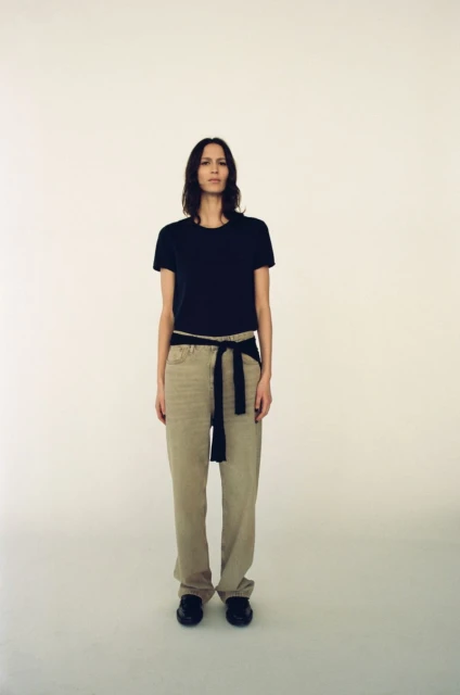Full-length studio shot of a woman in black T-shirt, beige pants cinched with a black sash, standing against a plain white background.
