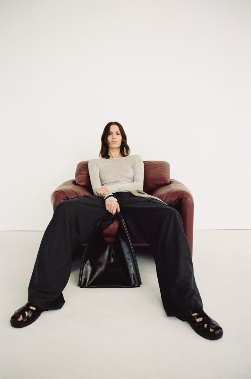 A woman with shoulder-length brown hair sits in a red leather chair with a black triangular handbag and wide-leg black pants against a white backdrop.