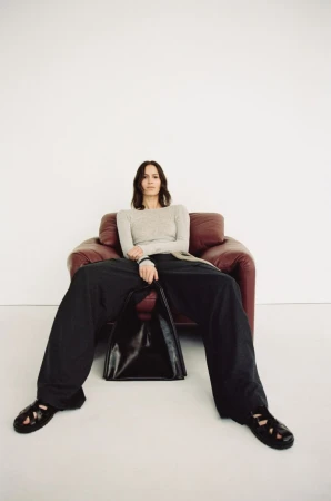 Jeanne Cadieu seated in a deep red leather armchair wearing wide-leg black trousers and holding a black tote against a white backdrop.