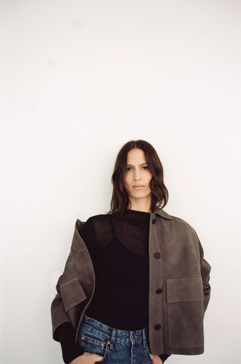 Woman in casual fashion poses against a white wall in a studio with natural light, wearing a jacket, top, and jeans for a fashion editorial.