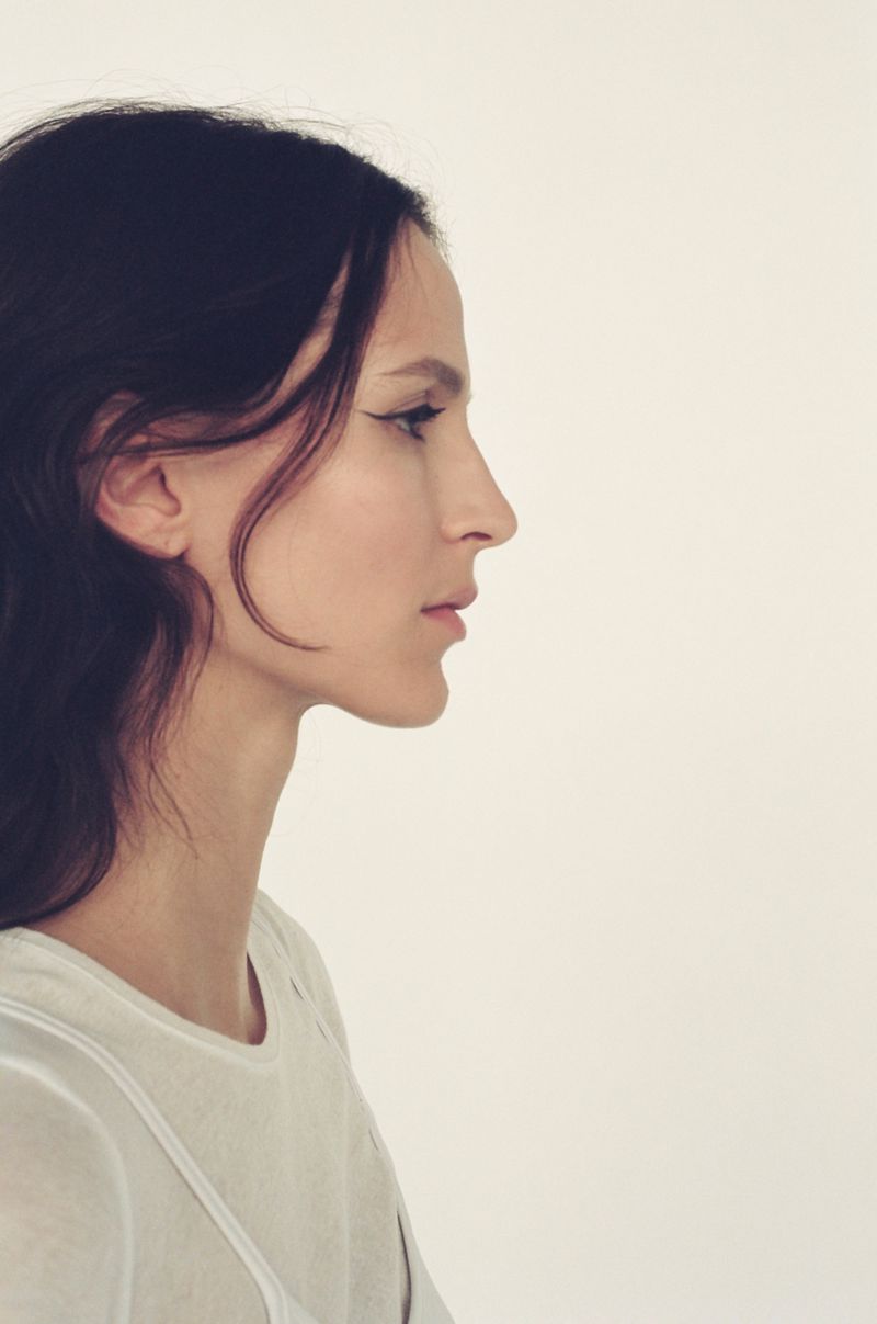 Profile portrait of a woman with dark hair, wearing a white top, against a light background. Beauty and fashion aesthetic.