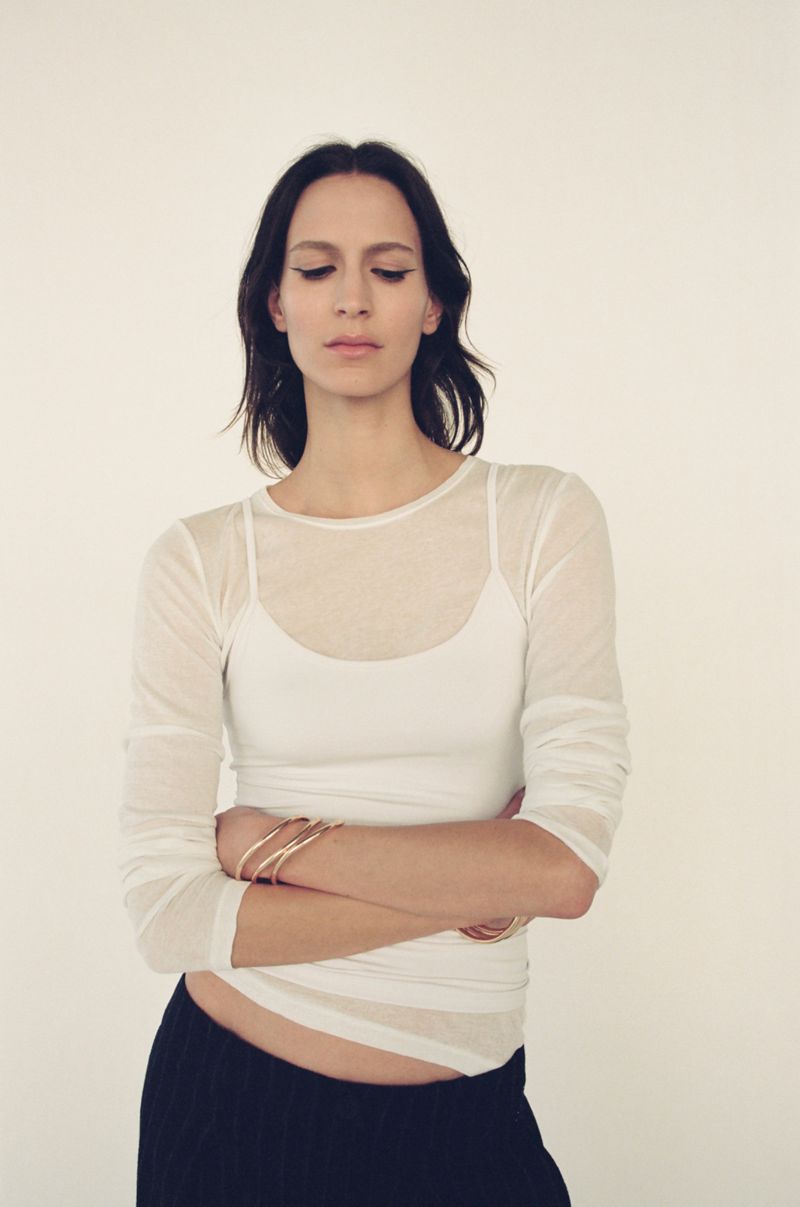 Woman with arms crossed, wearing a long-sleeved top and dark pants, looks downward in a brightly lit indoor space with a neutral background.