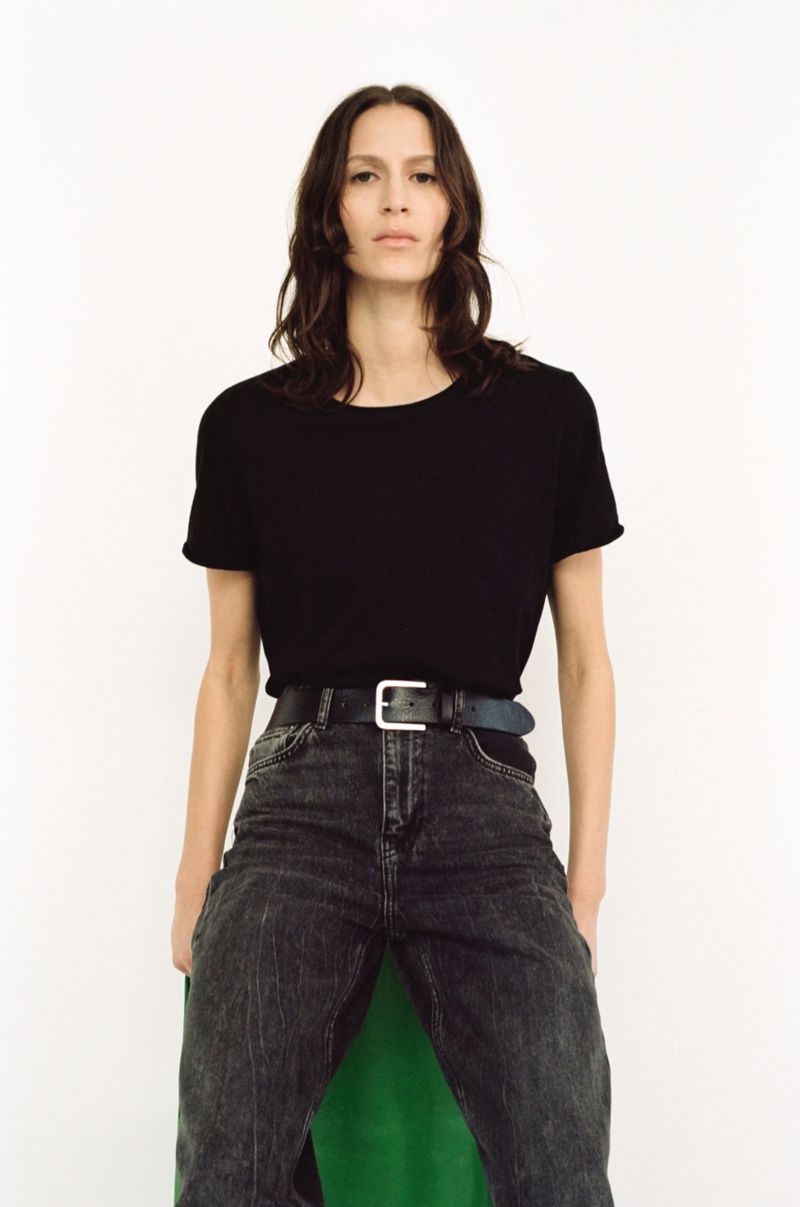 A woman stands in a photo studio wearing a black T-shirt, acid-wash jeans, and a silver-buckled belt against a stark white backdrop.
