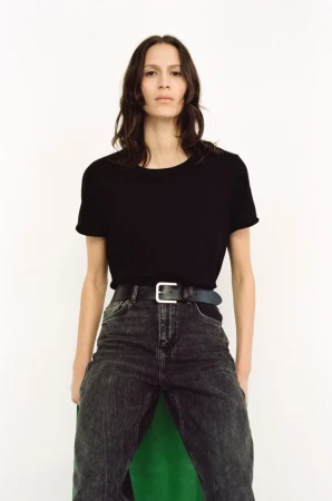 A woman stands in a photo studio wearing a black T-shirt, acid-wash jeans, and a silver-buckled belt against a stark white backdrop.