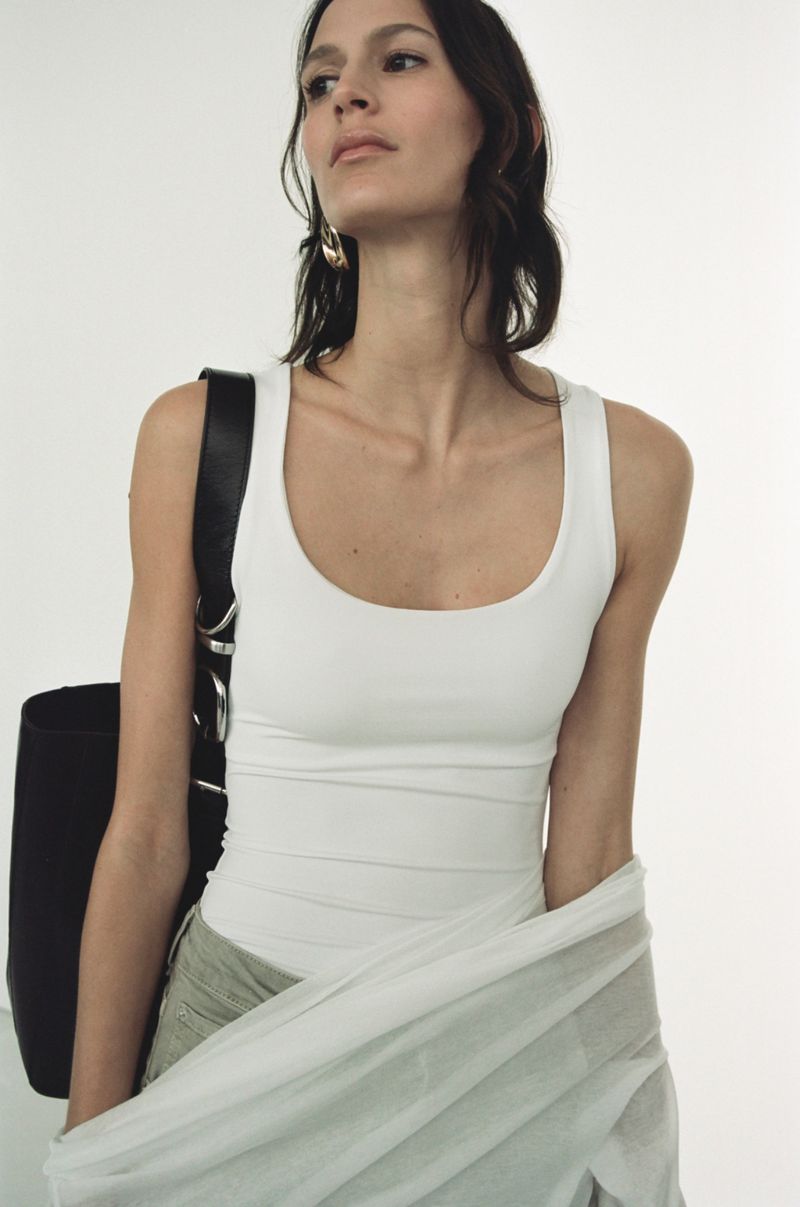 Woman in white tank top with black bag and light green skirt stands against a bright white background in a studio setting.