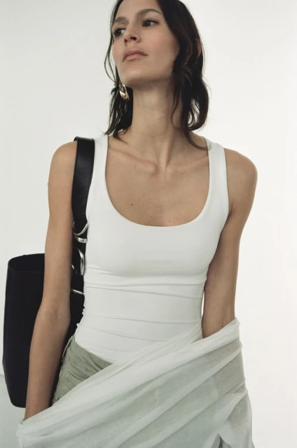 Woman in white tank top with black bag and light green skirt stands against a bright white background in a studio setting.
