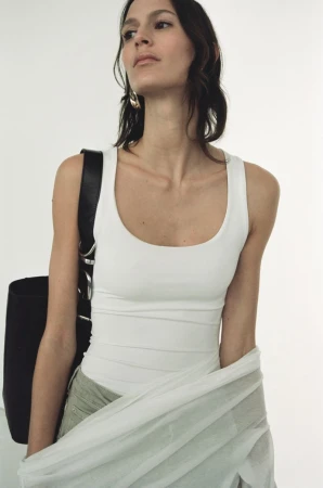 Woman in white tank top with black bag and light green skirt stands against a bright white background in a studio setting.