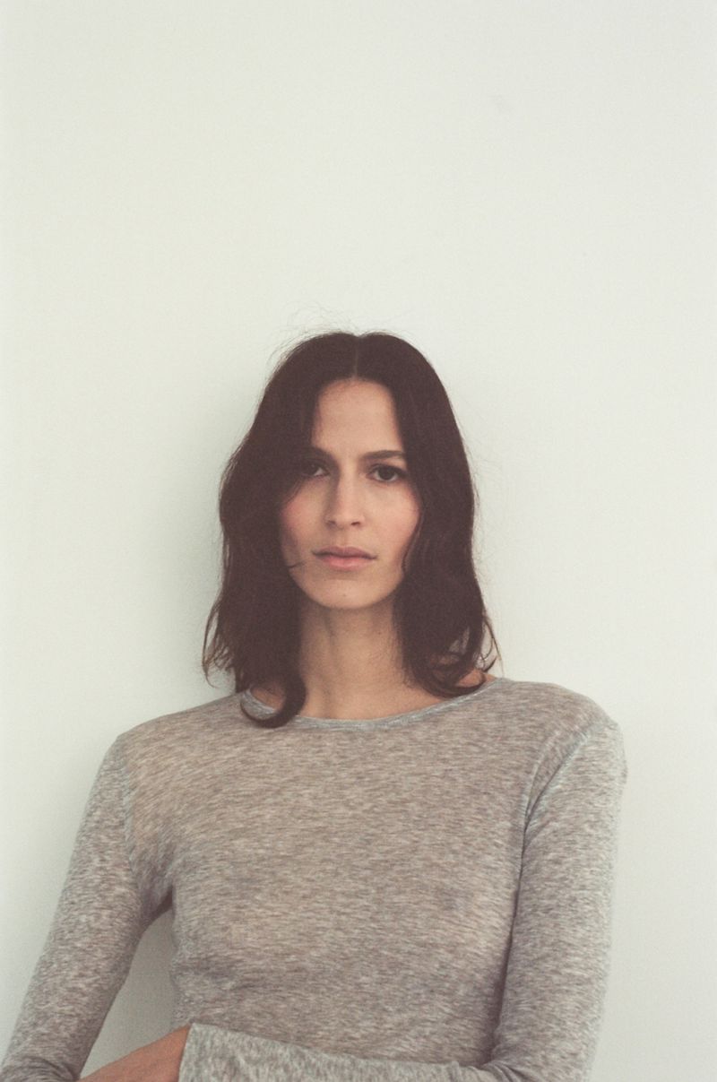 Portrait of an adult female with long brown hair wearing a grey long-sleeved shirt against a plain white background in a studio setting.