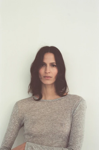 Portrait of an adult female with long brown hair wearing a grey long-sleeved shirt against a plain white background in a studio setting.