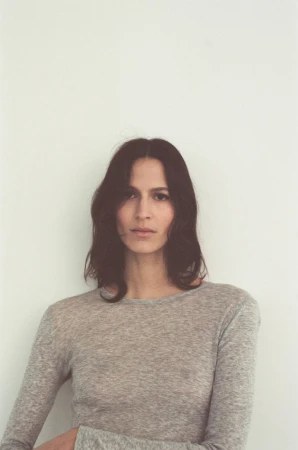Portrait of an adult female with long brown hair wearing a grey long-sleeved shirt against a plain white background in a studio setting.