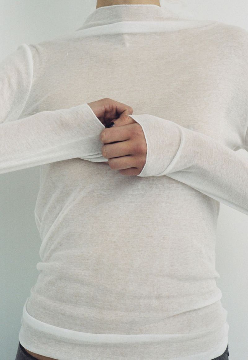 A person wearing a white long-sleeved shirt is shown indoors against a white background. The individual is shown from the chest up.