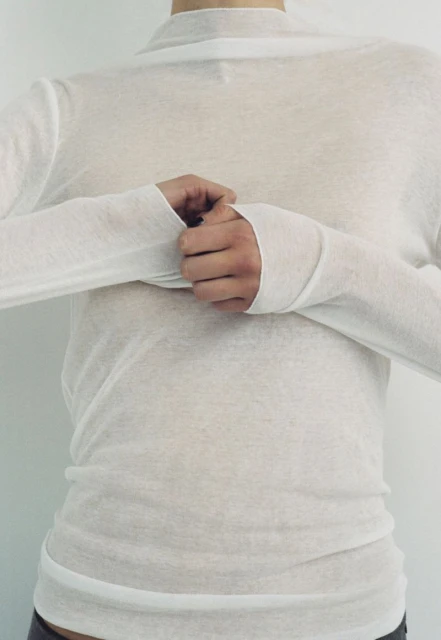 A person wearing a white long-sleeved shirt is shown indoors against a white background. The individual is shown from the chest up.
