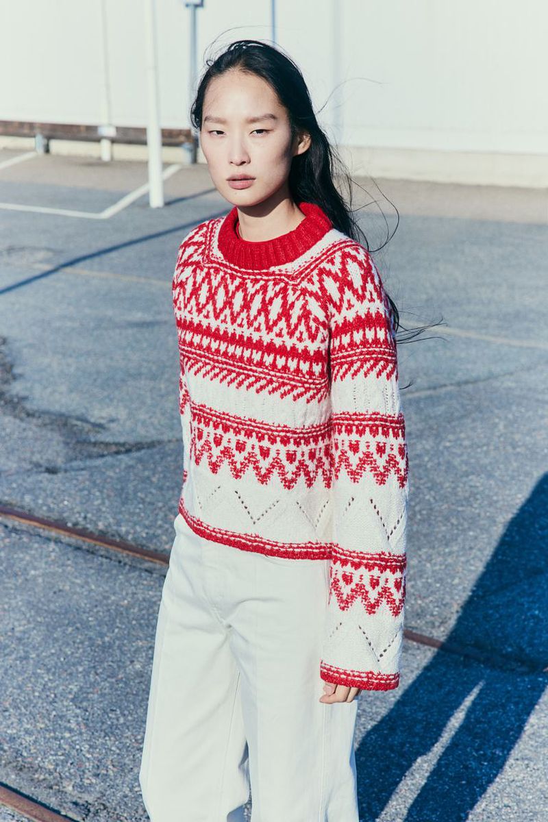 A woman with dark hair wears a red and white patterned sweater and white pants, posing in an outdoor urban environment. Fashion portrait.