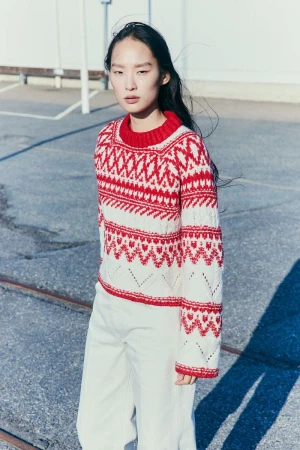 A woman with dark hair wears a red and white patterned sweater and white pants, posing in an outdoor urban environment. Fashion portrait.