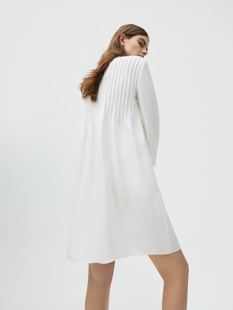 Back view of an adult woman posing indoors, wearing a white long-sleeved dress with pleated design detail under bright studio lighting.
