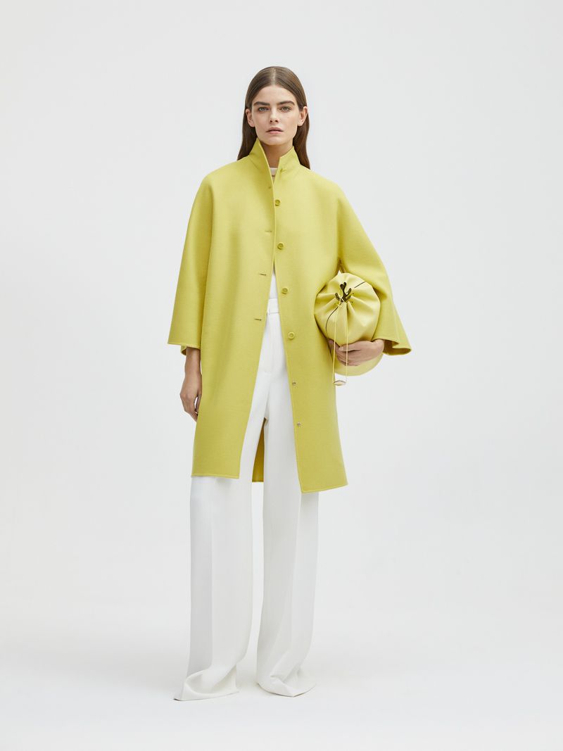 Woman in a chartreuse coat and white pants holding a chartreuse pouch bag in a brightly lit photo studio against a white backdrop.