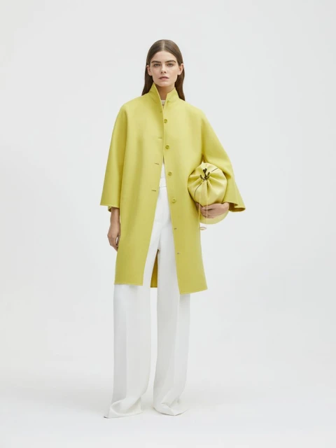 Woman in a chartreuse coat and white pants holding a chartreuse pouch bag in a brightly lit photo studio against a white backdrop.