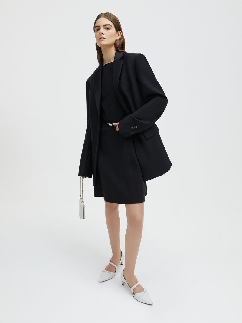 A stylish adult woman in a black blazer and dress stands confidently in a studio, accessorized with white heels and a small handbag.