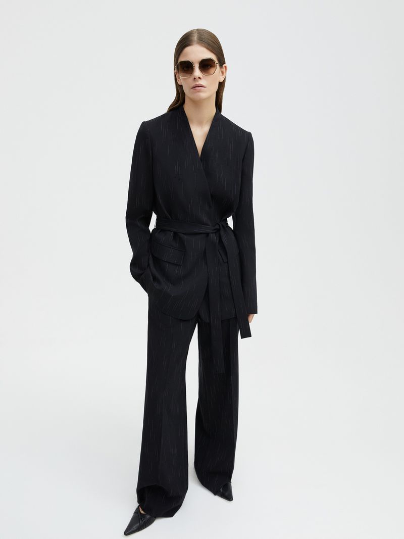 Woman in a black pinstripe suit with sunglasses stands in a photo studio against a white backdrop. Fashion editorial shot.