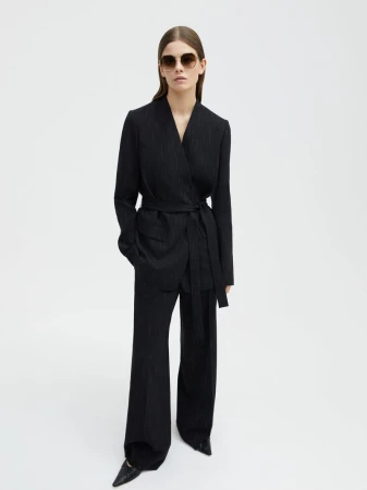 Woman in a black pinstripe suit with sunglasses stands in a photo studio against a white backdrop. Fashion editorial shot.