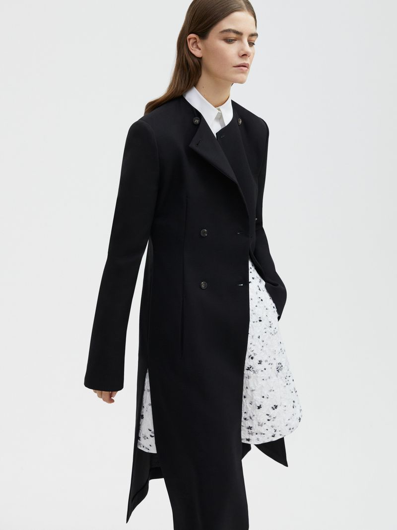 A woman in a black coat and white blouse poses in a brightly lit studio against a white background, showcasing modern fashion.