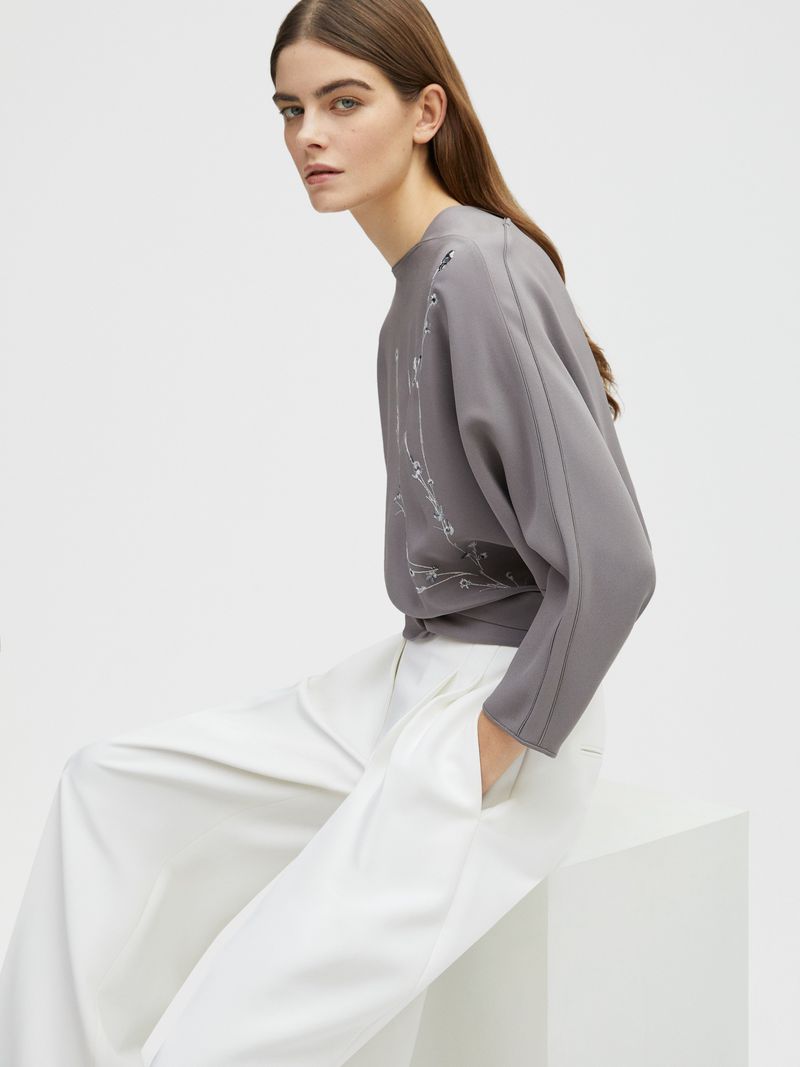 A woman poses indoors, wearing a grey top with embroidery and white pants, seated on a white cube, against a clean white backdrop.