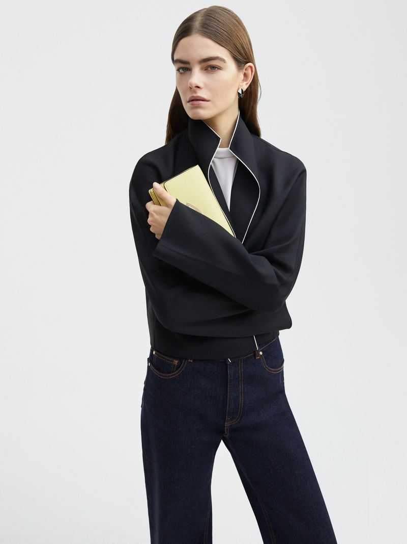 Model Merlijne Schorren wearing a black structured jacket and dark denim jeans holding a yellow clutch against a plain background.