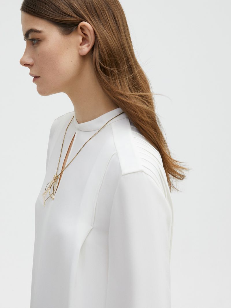 Woman in white top with gold necklace against a neutral background. Minimalist aesthetic with focus on fashion and portraiture.