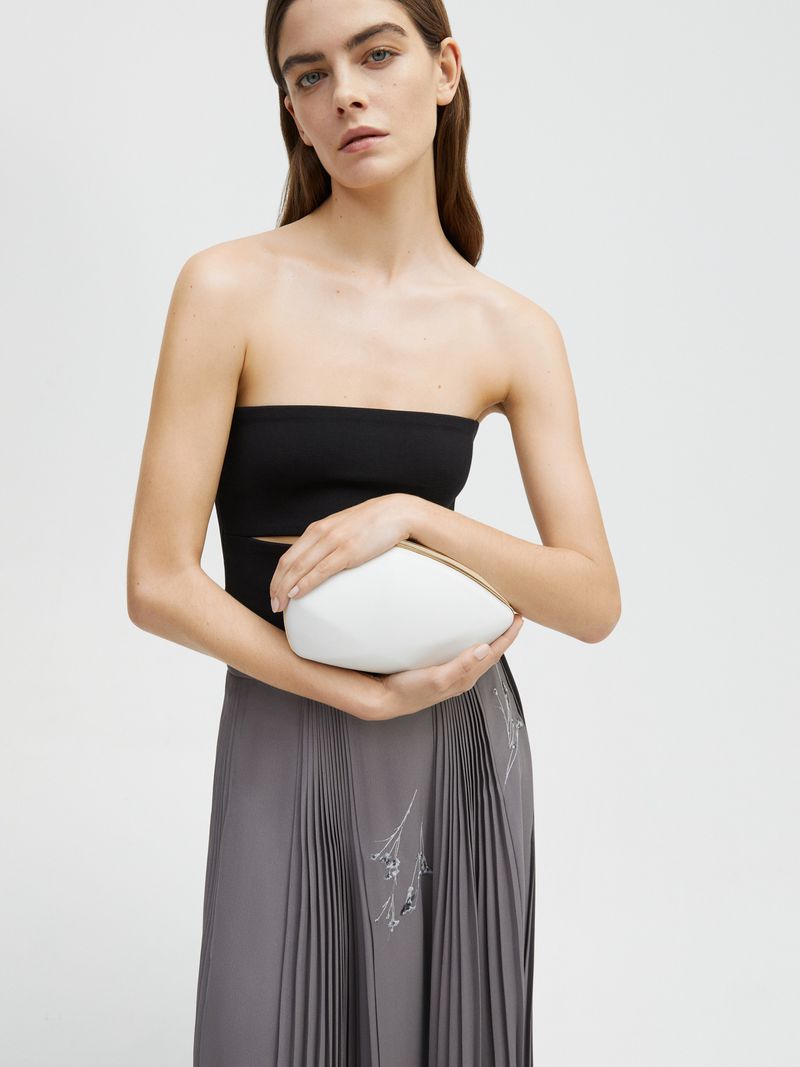 A model poses in a black top and grey skirt, holding a unique white clutch in a studio setting, emphasizing fashion and style.