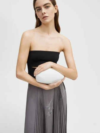 A model poses in a black top and grey skirt, holding a unique white clutch in a studio setting, emphasizing fashion and style.