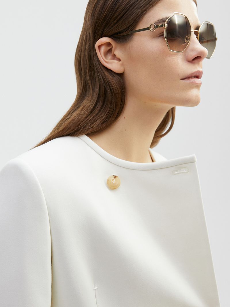 Woman in white coat wearing sunglasses, close-up portrait. Fashion shot in a bright indoor setting with natural-looking soft light.