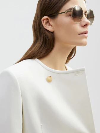 Woman in white coat wearing sunglasses, close-up portrait. Fashion shot in a bright indoor setting with natural-looking soft light.