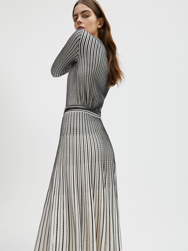 A woman poses in a black-and-white striped, long-sleeved dress and skirt in a studio setting, looking toward the camera.