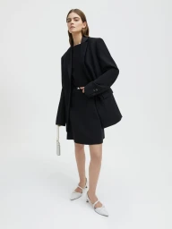 Model Merlijne Schorren wearing a black oversized blazer and matching black dress with white slingback heels against a white studio backdrop.