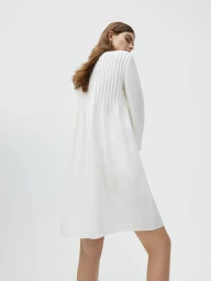 Model Merlijne Schorren seen from behind wearing a white pleated long-sleeve dress against a seamless studio backdrop.