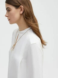 Profile view of a female model wearing a white top and a long gold necklace against a plain white background.