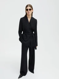 Model wearing a tailored black belted pinstripe suit and sunglasses against a plain white studio backdrop.