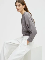 Model seated on a white cube wearing a grey top and white trousers against a white seamless background