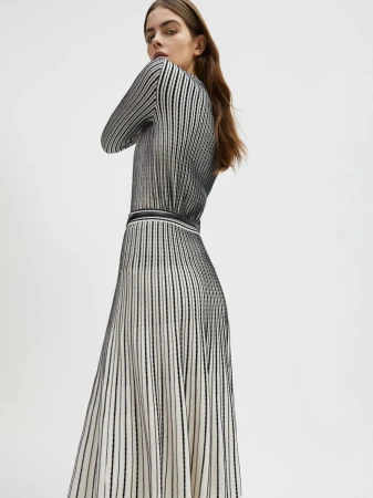 Model Merlijne Schorren wearing a black and white vertically striped long knit dress against a plain white studio background.