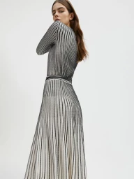 Model Merlijne Schorren wearing a black and white vertically striped long knit dress against a plain white studio background.