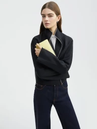 Model Merlijne Schorren wearing a black structured jacket and dark denim jeans holding a yellow clutch against a plain background.