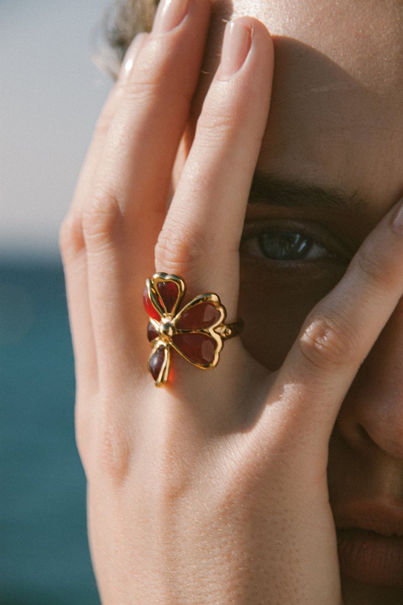 Close-up of a woman's hand with a red and gold flower ring against a blue sky and ocean background, showcasing jewelry details.