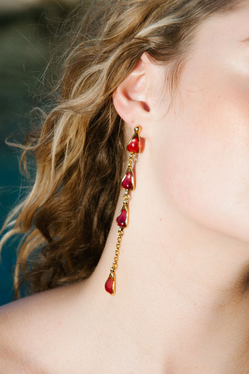 Close-up of a woman with blonde curly hair wearing long, ornate gold earrings with red teardrop gemstones against a blurry teal background.