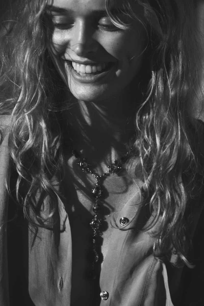 Close-up black and white shot of a smiling woman with curly hair wearing a necklace and buttoned shirt, with dramatic lighting.