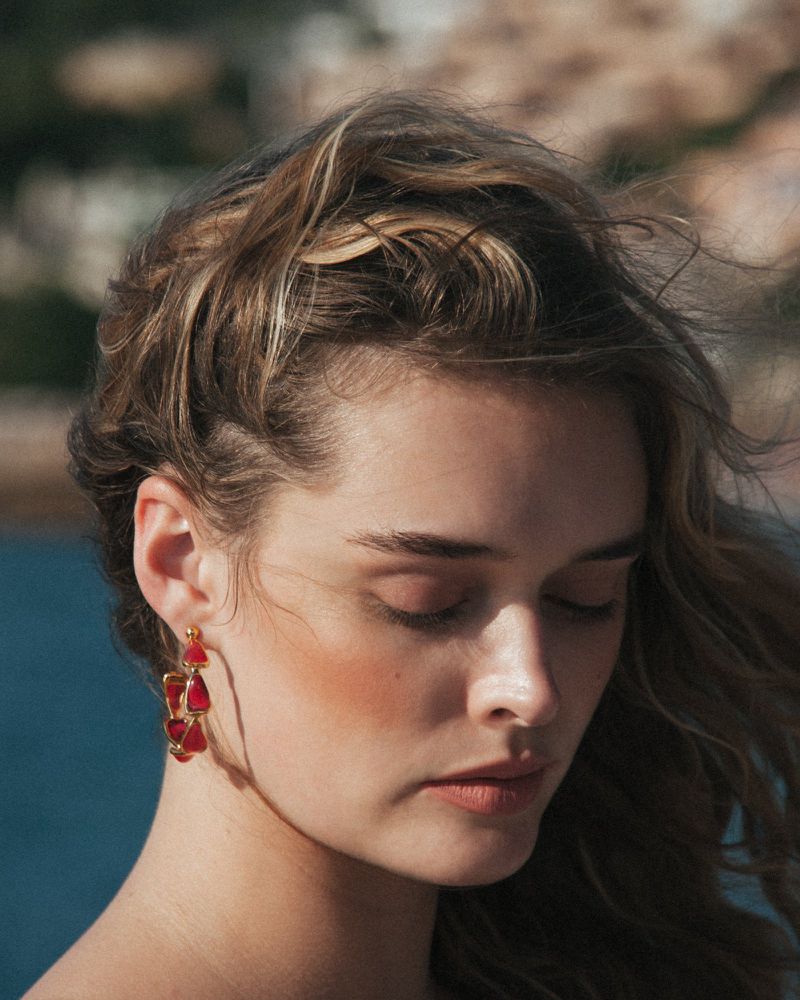 Close-up of a woman with light brown hair wearing red drop earrings, eyes closed, against a blurred backdrop of sea and vegetation.