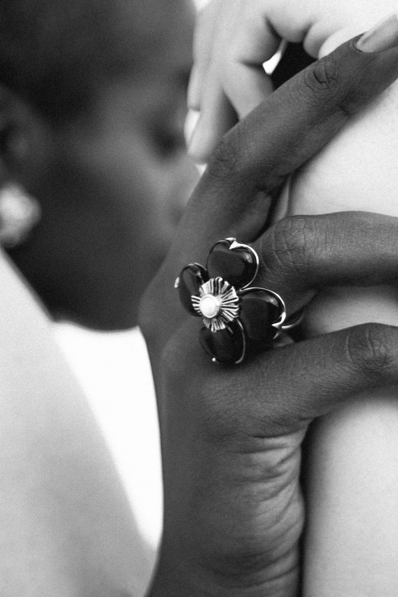Monochrome closeup of two people holding hands, one person wearing a flower ring, in an indoor setting.