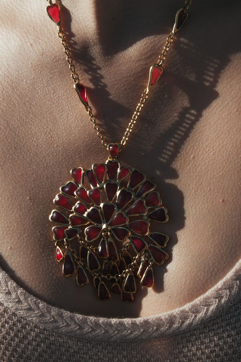 Close-up of a gold necklace with a large, round red pendant. The necklace sits on a person's chest, with shadows and highlights.